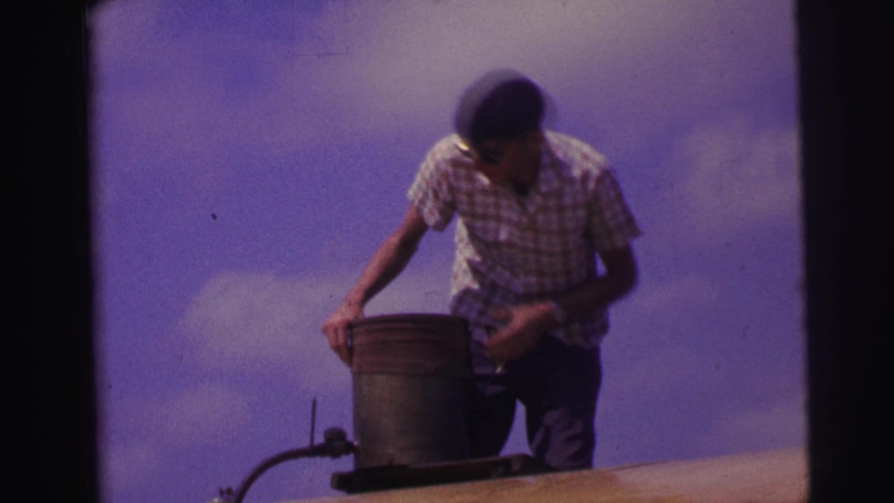 Man in plaid shirt and sunglasses stands by a metal bucket on a rooftop.