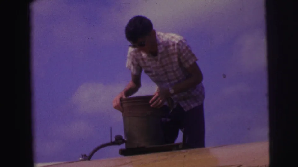 A man in plaid shirt and sunglasses bends over a metal rooftop container in vintage 8mm footage from Brazil.