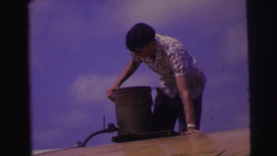 The man reaches one hand across the rooftop while steadying himself near a metal bucket and small pipe or hose fitting.