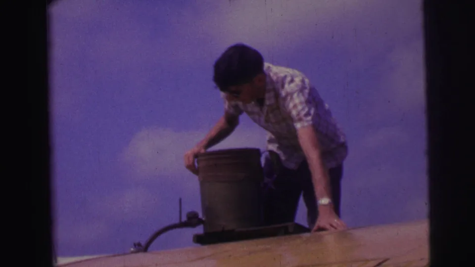 A man in sunglasses steadies himself with one hand on the roof while standing beside a metal container in the 1967 Brazil reel.