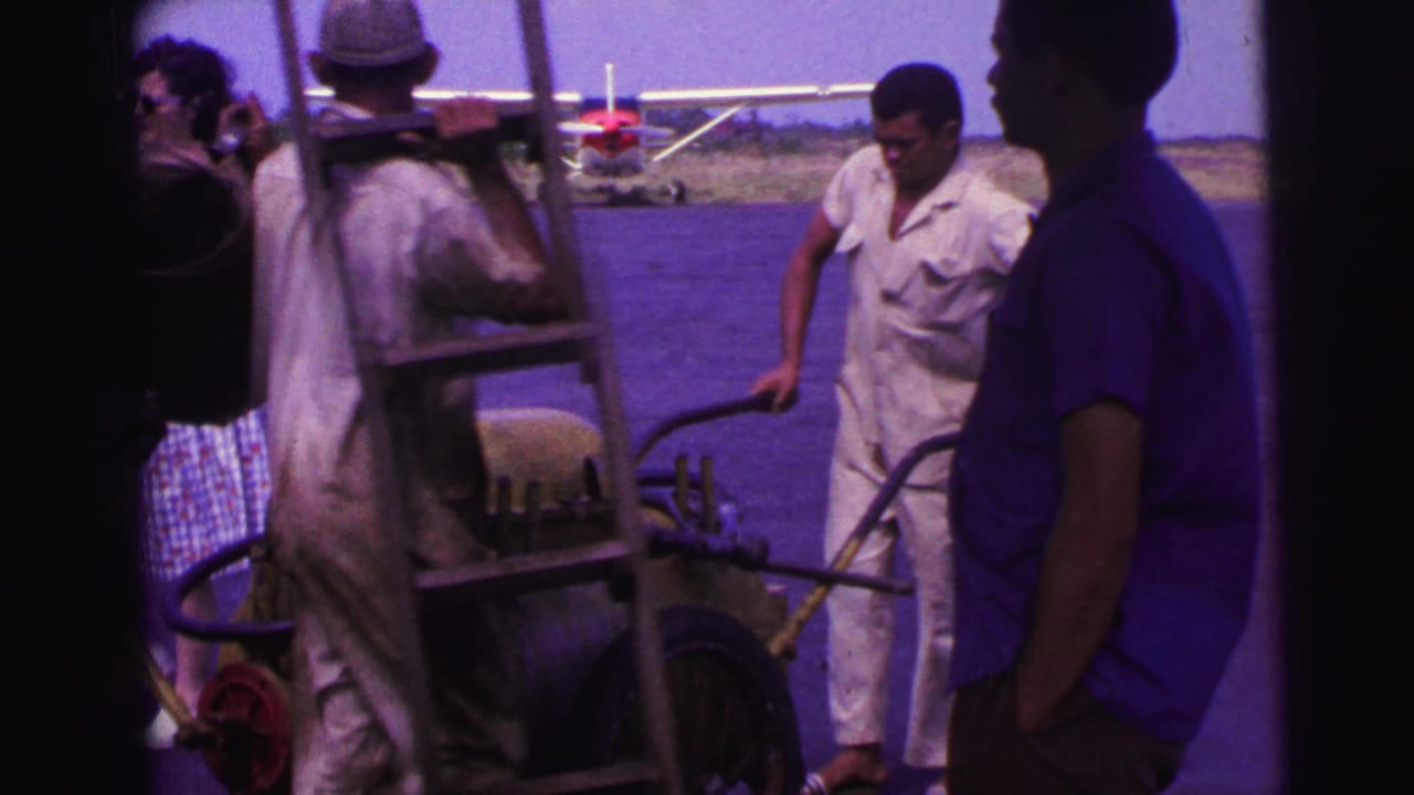 1967 Brazil home-movie frame showing workers near a yellow fuel cart, ladder, and small plane on a tarmac.