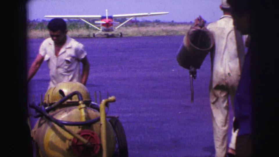 1967 Brazil home-movie frame showing a ground worker beside a yellow fuel cart and another carrying equipment near a small plane.