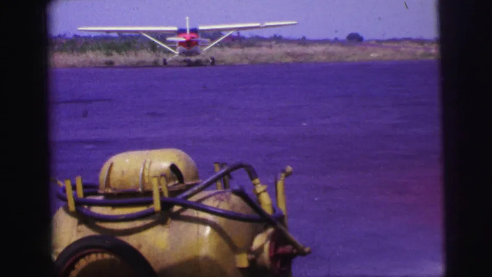 1967 Brazil airfield frame focused on a yellow fuel cart and hoses, with a red-and-white single-engine plane in the distance.