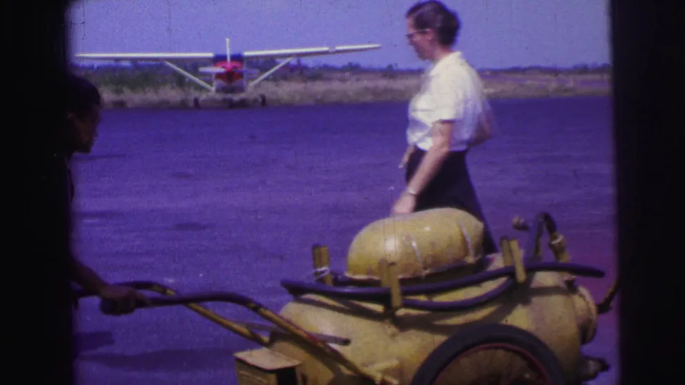 A woman in a white blouse and dark skirt walks beside the yellow fuel cart, with a small seaplane visible behind her in 1967 Brazil footage.