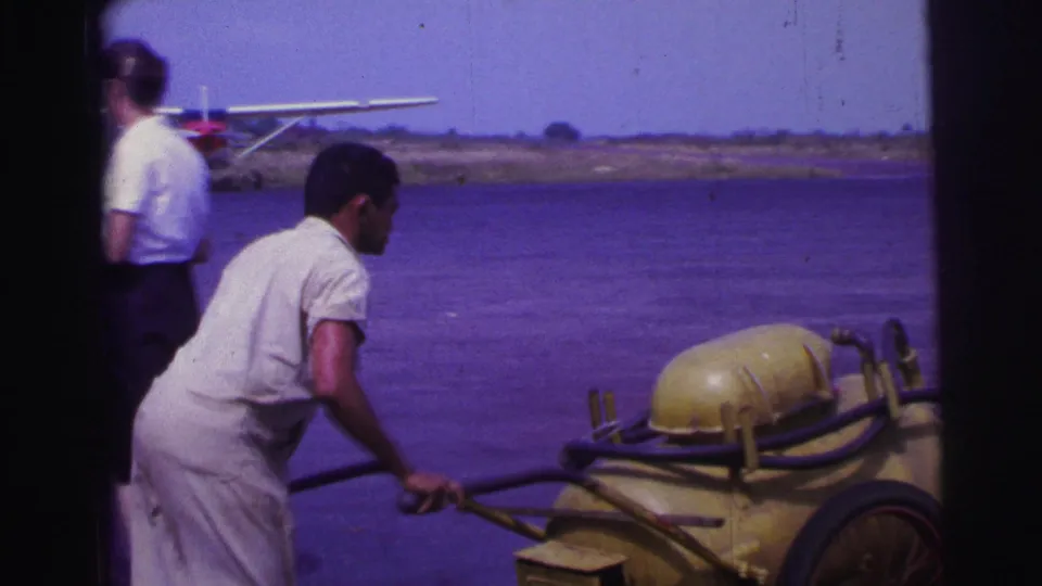 A man in light clothing handles the yellow fuel cart as a woman walks nearby and a red and white seaplane sits on the tarmac in Brazil.