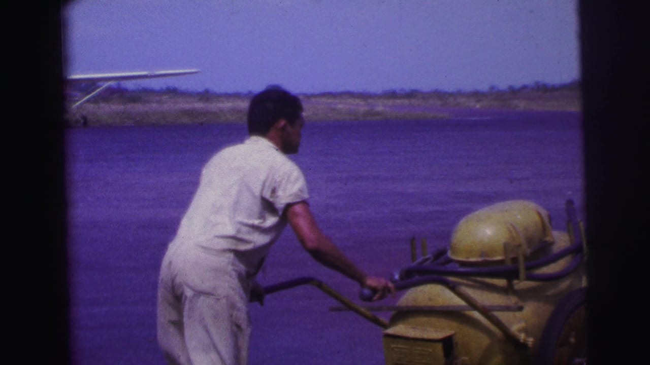 Man in a white jumpsuit pushes a yellow fuel cart near water or open airfield ground.