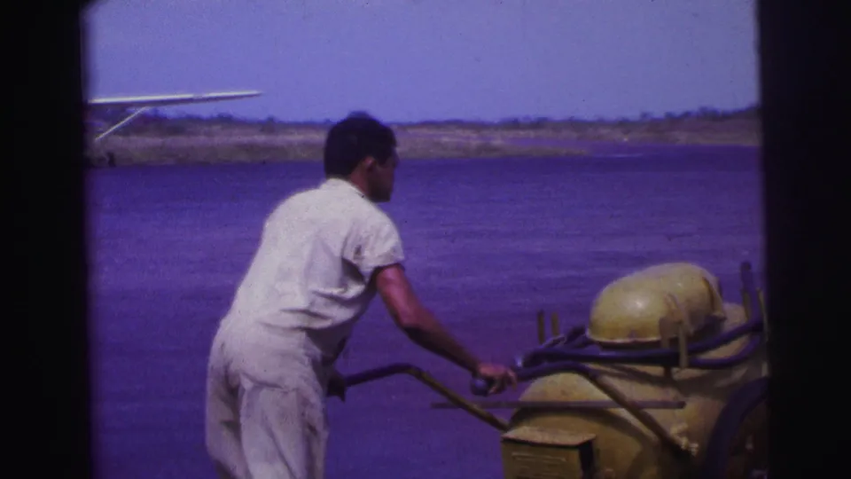 1967 Brazil home-movie frame showing a man in white work clothing hauling a yellow fuel cart across an airfield area.