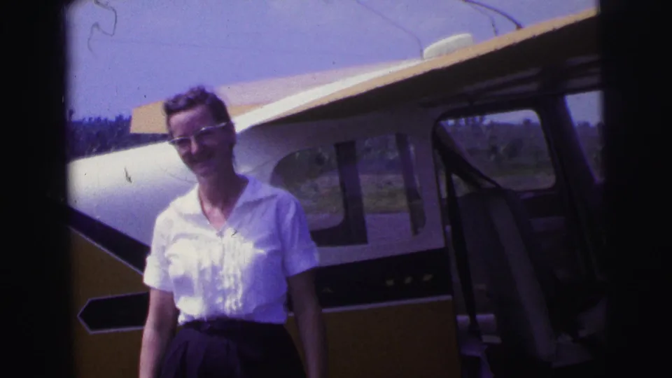 1967 Brazil home-movie frame showing a woman in a white blouse and dark skirt standing near a small airplane.
