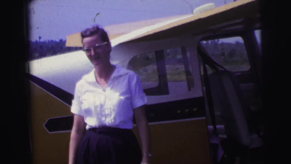 1967 Brazil 8mm frame of a woman in glasses beside a small aircraft, smiling or speaking toward the camera.