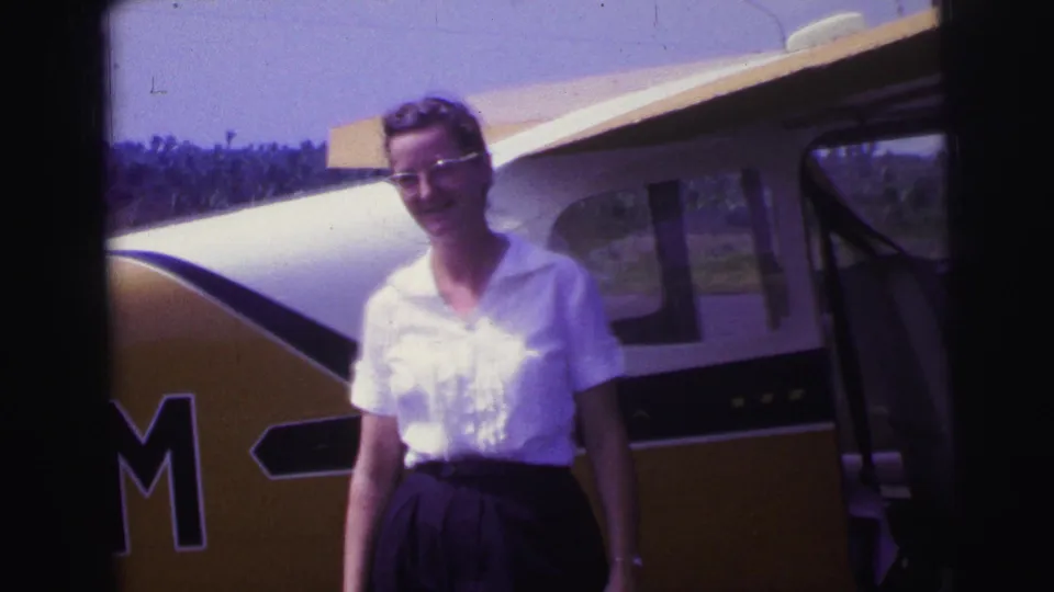A smiling woman in glasses stands beside the small aircraft, with its open door and partial lettering visible.