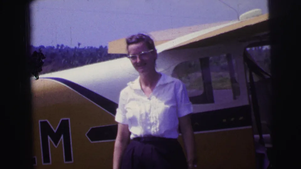 The woman remains posed near the yellow-and-white aircraft, with partial side lettering and a tree line behind her.