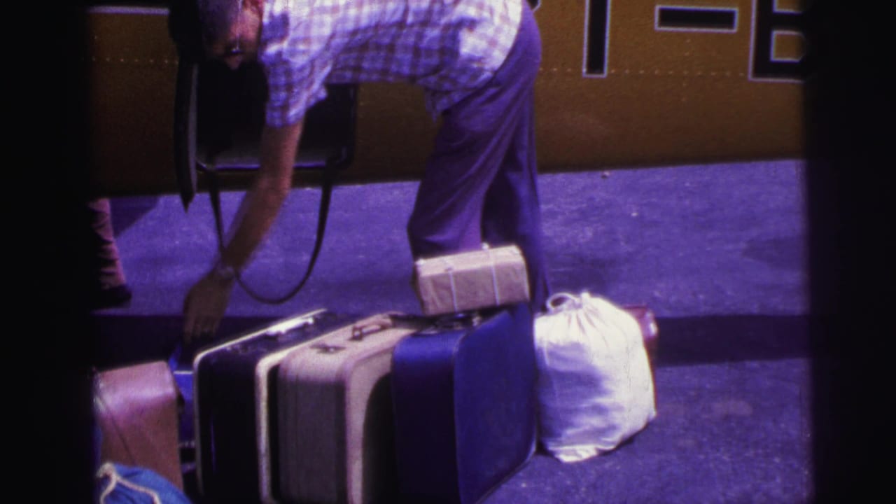 1967 Brazil home-movie frame of a man bending by stacked luggage beside the open door of a yellow aircraft with partial markings.