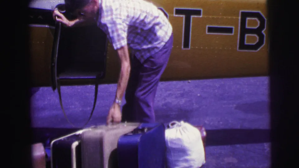 1967 Brazil home-movie frame of a man adjusting suitcases beside a yellow aircraft marked with partial T-B lettering.