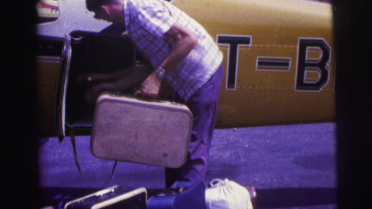 Man bends beside a small yellow-and-white airplane while holding a hard suitcase near an open compartment.