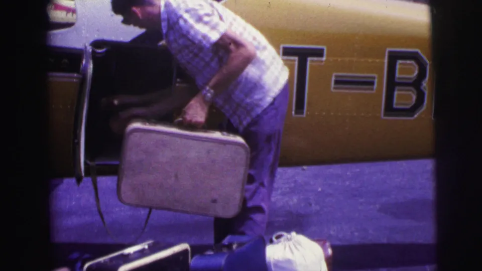 In 1967 Brazil footage, a man in a checked shirt loads a hard suitcase beside a yellow-and-white small airplane on a tarmac.