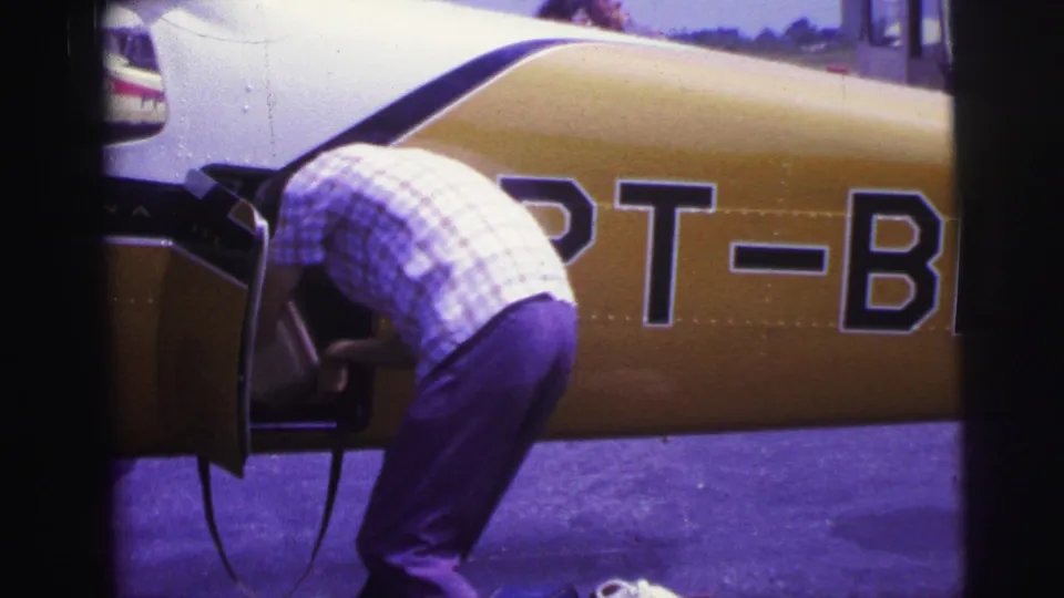 A man bends into a small yellow-and-white aircraft compartment on a Brazilian tarmac, with partial PT-B lettering visible on the fuselage.