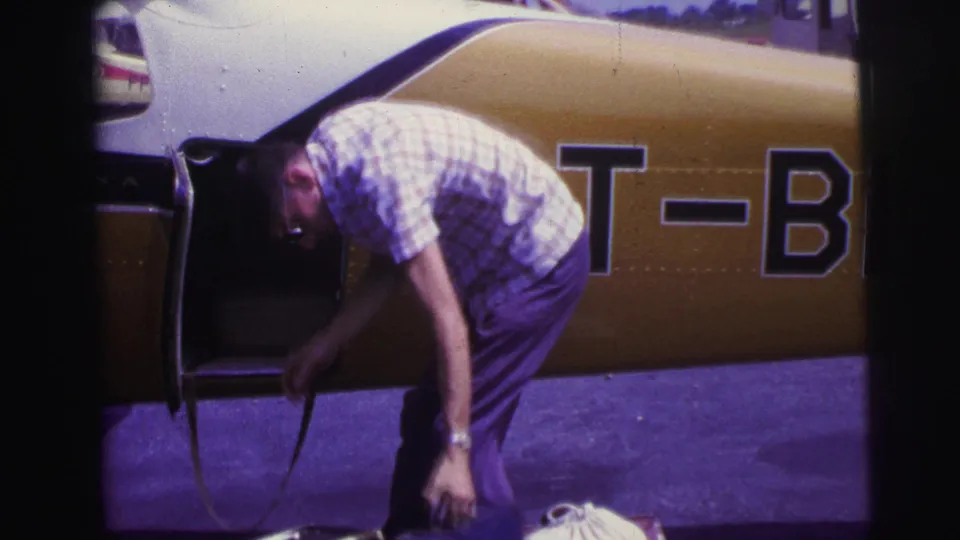 In 1967 Brazil home-movie footage, a man in a plaid shirt bends near the open side of a yellow and white small plane on an outdoor airfield.
