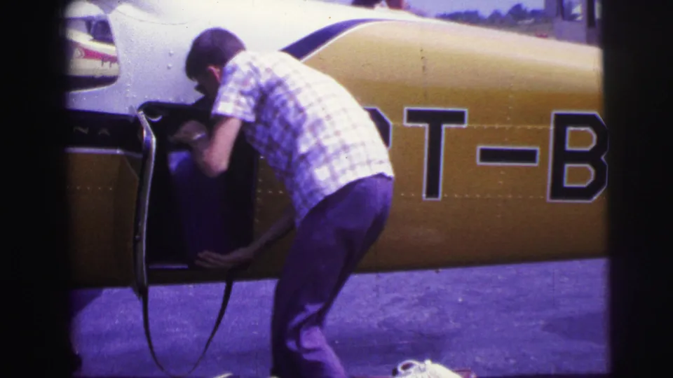 A man in a plaid shirt places a blue suitcase into the open side compartment of a small yellow and white plane, with registration letters partly visible.