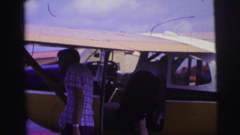 The man stands beside the small plane under its wing, with the open cabin area, propeller, struts, and another person partly visible nearby.