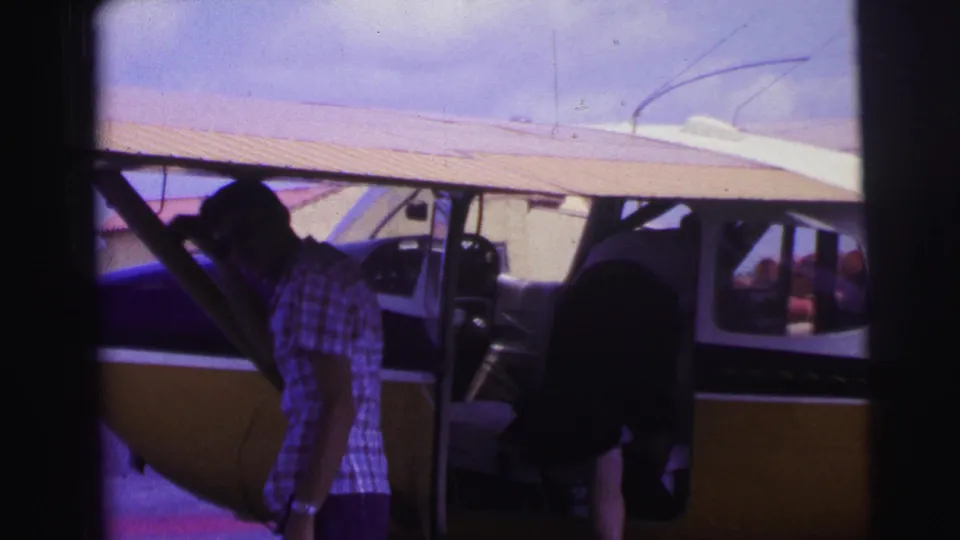 In 1967 Brazil home-movie footage, a man stands by the open door of a yellow and blue single-engine aircraft at an airfield.