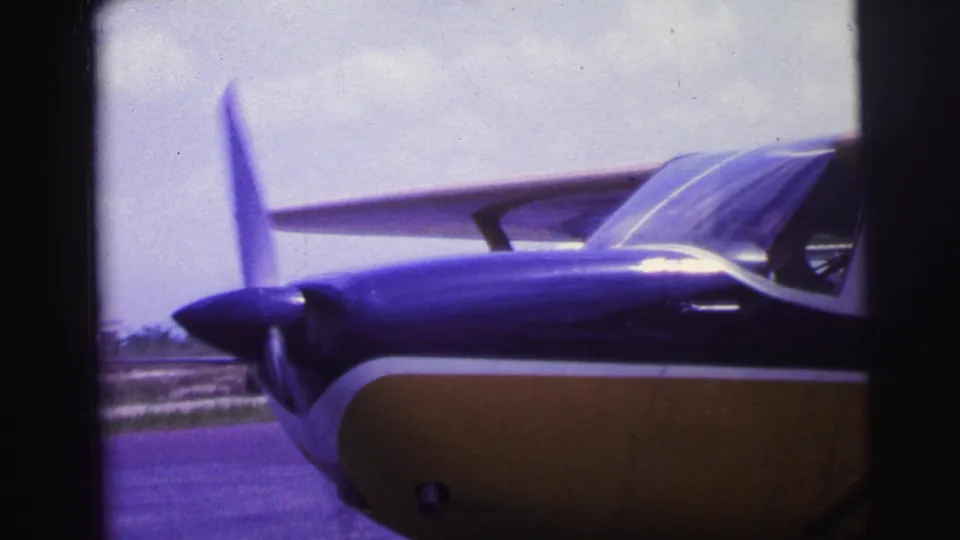The camera frames the nose, propeller, wing, and cockpit windows of the blue and yellow aircraft on the airfield.