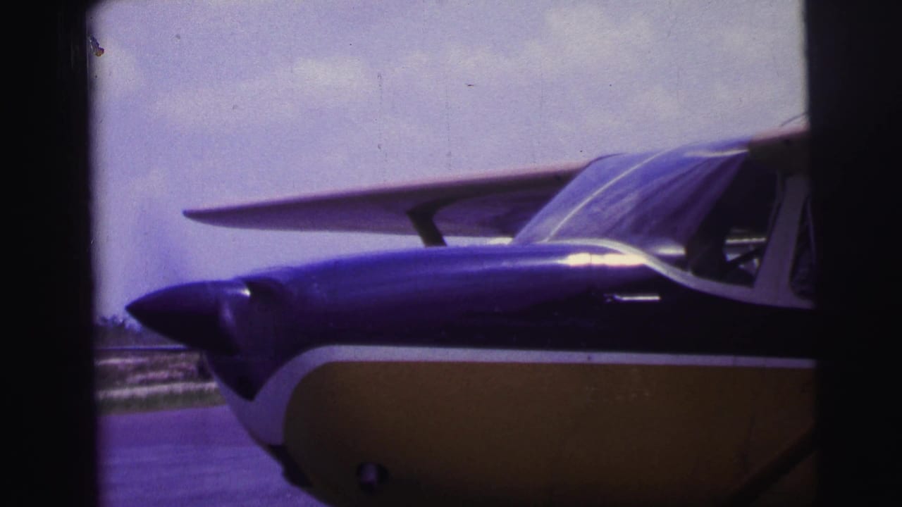 1967 Brazil 8mm frame showing the nose and cockpit area of a small blue-and-yellow propeller airplane at an airfield.