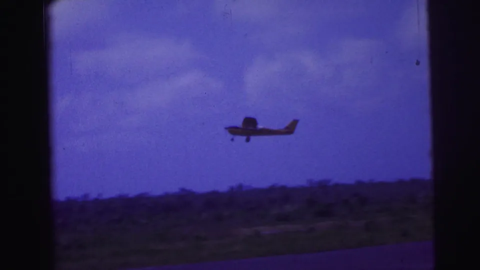 1967 home-movie frame of a small aircraft climbing above a low Brazil airfield landscape.
