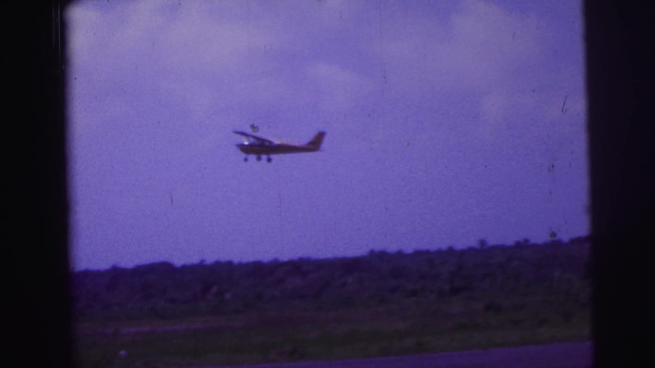 1967 Brazil 8mm frame showing a small propeller aircraft just after takeoff above a rural horizon and purple-tinted sky.