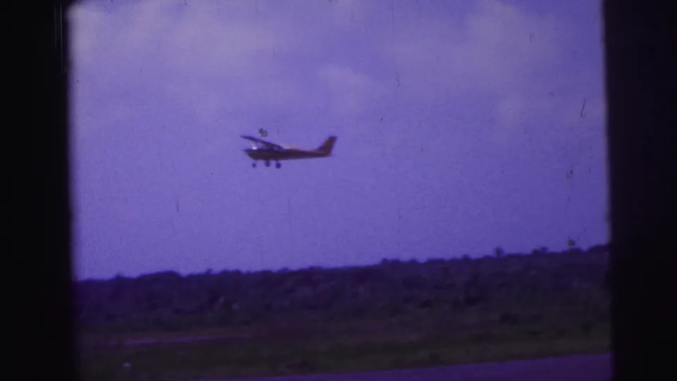 1967 Brazil 8mm frame showing a small propeller aircraft just after takeoff above a rural horizon and purple-tinted sky.