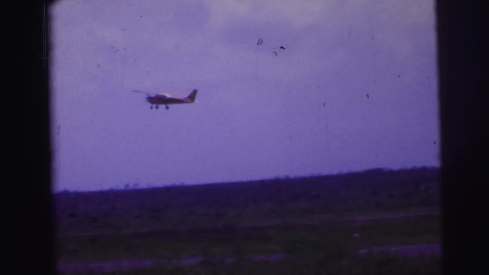 1967 Brazil 8mm view of a small aircraft crossing the sky above a rural horizon, with dark vertical edges in the frame.