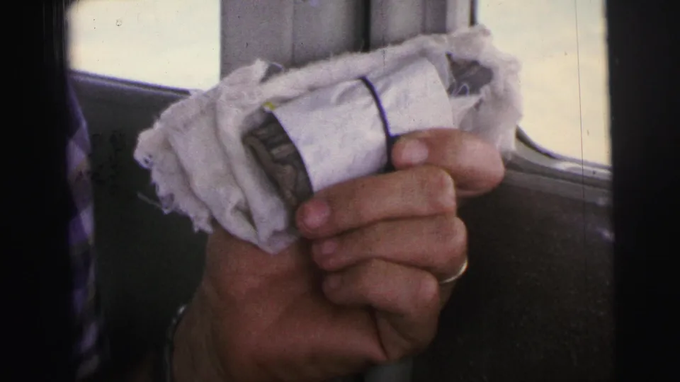 A close 8mm view shows a ringed hand holding a white bundled package beside an aircraft window in Brazil travel footage.