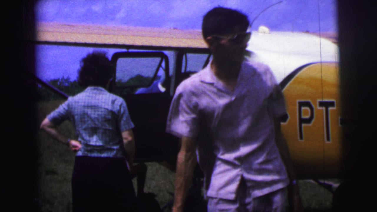 1967 Brazil home-movie frame showing people near a small airplane, with an open cabin door and PT letters visible on the yellow tail section.