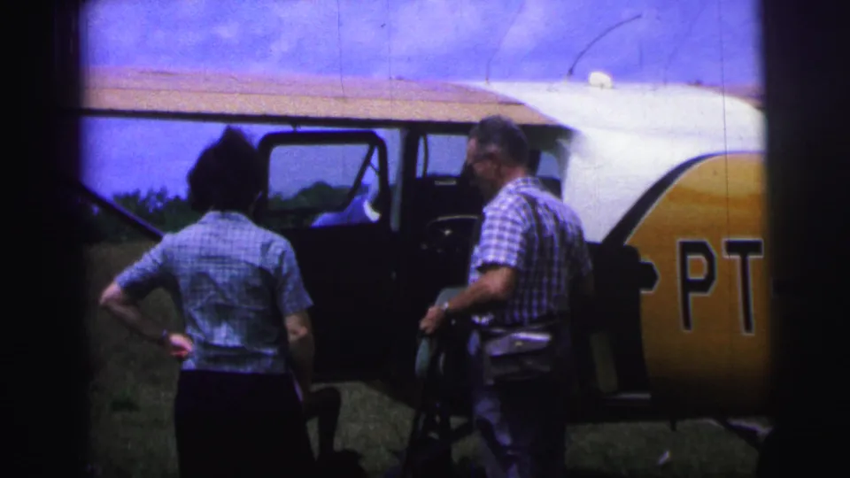1967 Brazil frame showing a woman and a man beside an open small-airplane door, with part of the yellow PT-marked tail visible.