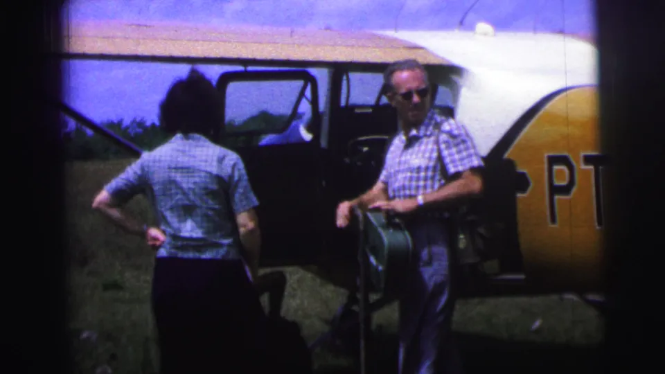 1967 Brazil home-movie frame of a man with sunglasses holding a green suitcase near a small airplane as a woman faces the open door.