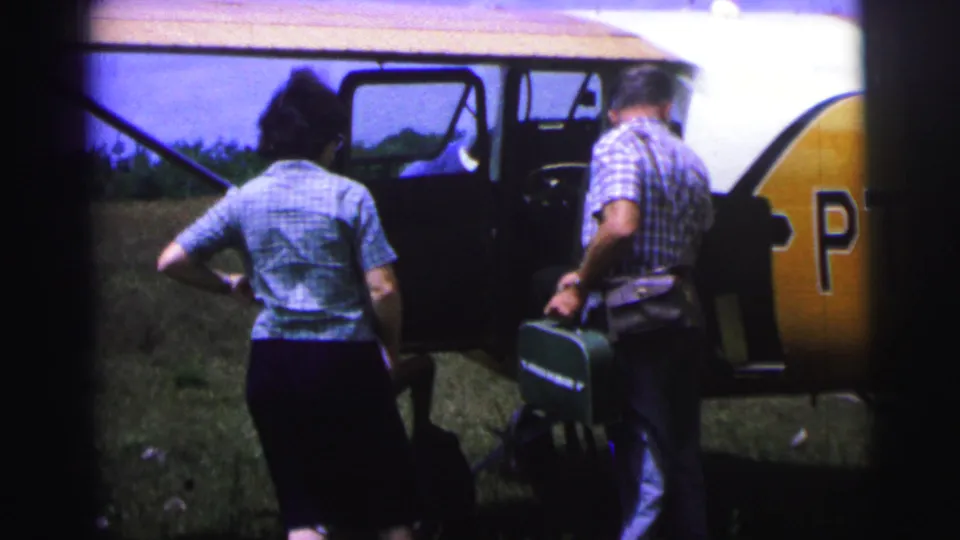 1967 Brazil frame showing a man carrying a green suitcase beside the open door of a small airplane while a woman stands facing the aircraft.