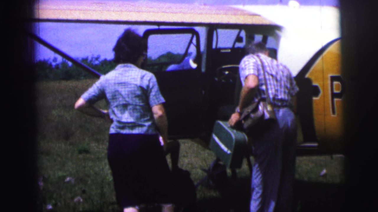 In 1967 Brazil home-movie footage, two adults stand near a small aircraft while a man carries a green suitcase across grass.