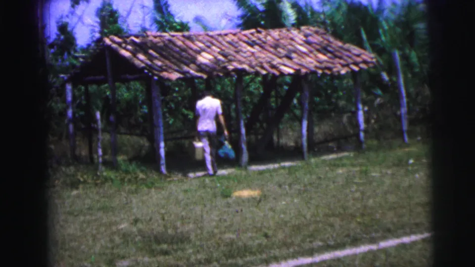 A traveler in the Brazil 1967 home movie walks with luggage toward a rustic tiled shelter near tropical vegetation.
