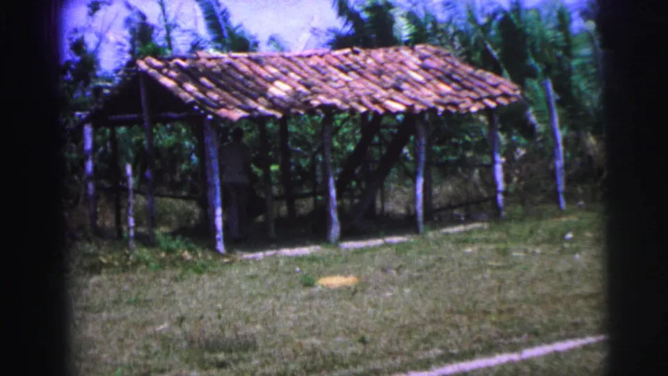 A rustic open shelter in 1967 Brazil stands amid grass and vegetation, with a person faintly visible in the shaded interior.