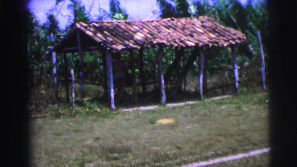 1967 Brazil 8mm frame showing a rustic open shelter with a tiled roof, wooden posts, grass, and surrounding trees.