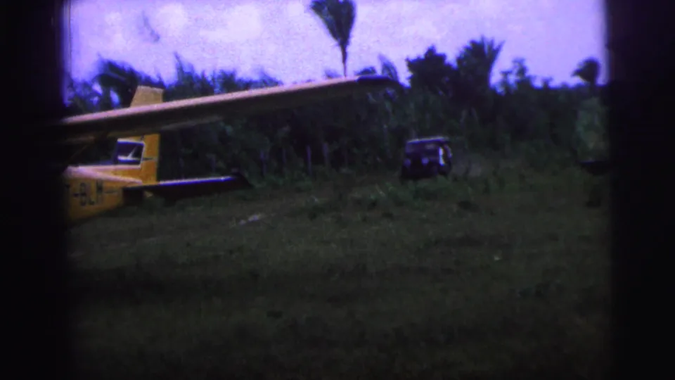 1967 Brazil 8mm frame showing a yellow single-engine plane in a grassy clearing with a jeep and palms behind it.