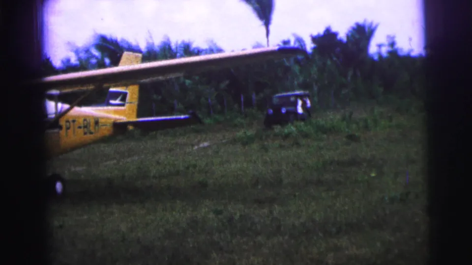 1967 Brazil 8mm frame of a yellow parked plane with visible tail markings, a jeep, tall grass, and palm trees.