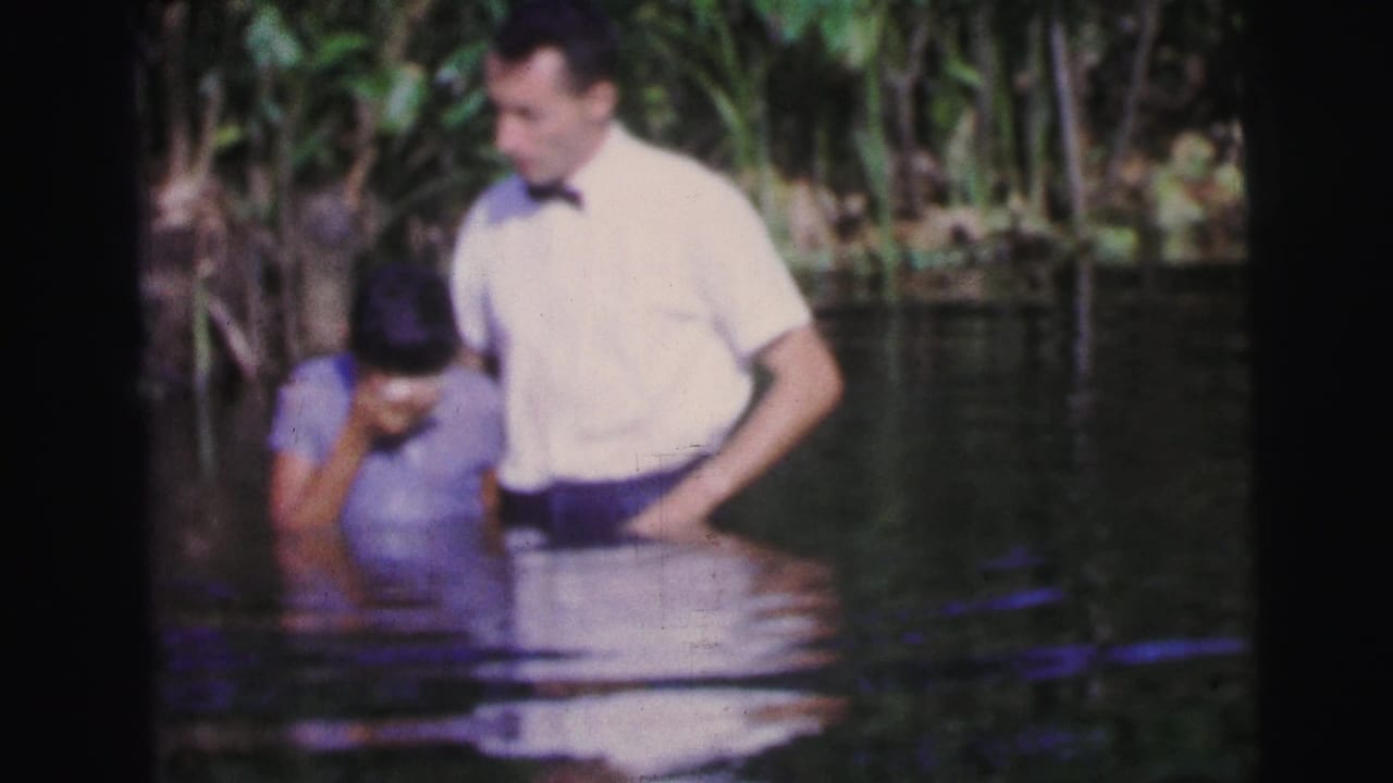 Man in a bow tie helps a person being lowered into river water.