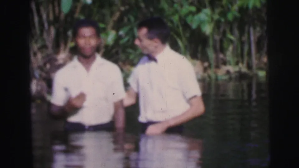 Two men stand in a tropical river in Brazil, one facing forward and one in a bow tie turned toward him.