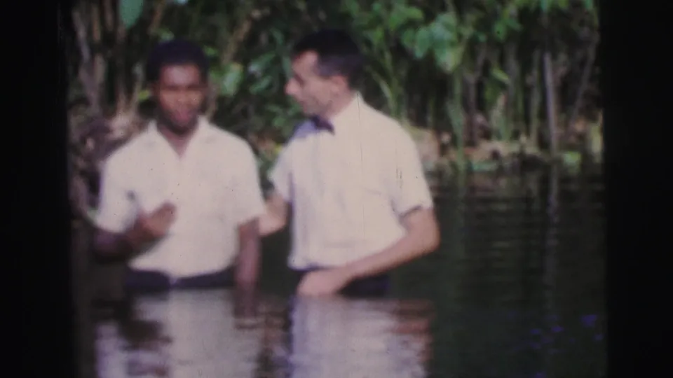 Two men in white shirts remain waist-deep in river water, with dense tropical foliage filling the background.