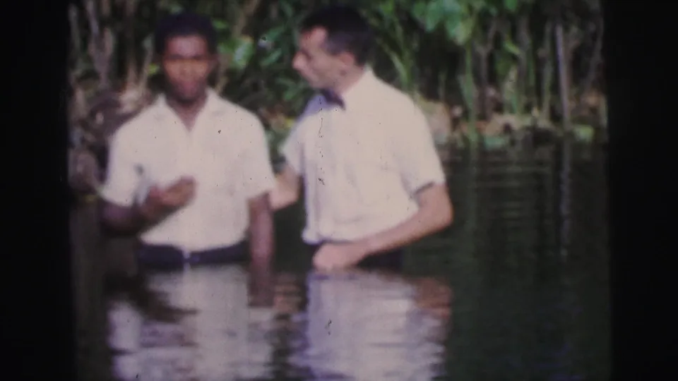 In the closing frame, two men stand waist-deep in a river, continuing a quiet exchange in the 1967 Brazil baptism footage.