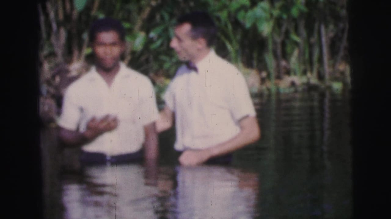 Two adult men in light shirts stand waist-deep in a tropical river in 1967 Brazil home-movie footage.