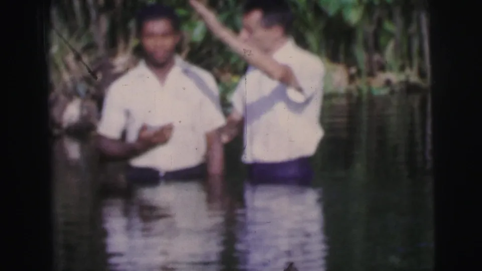 One man gestures over another as both stand waist-deep in a tropical river setting in 1967 Brazil footage.