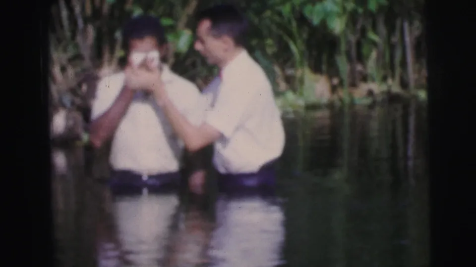 At the apparent ritual moment, one man touches the other's face or head as they stand in the river in Brazil.