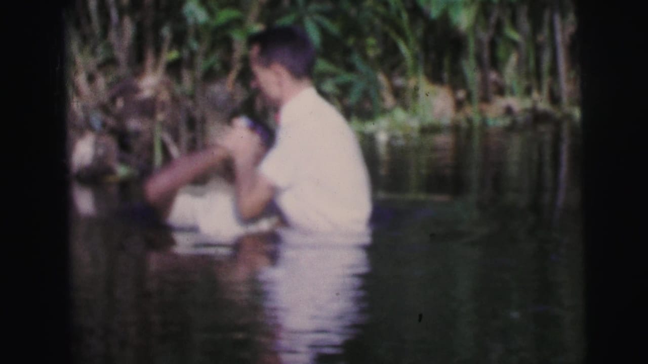 Two men in white shirts stand waist-deep in water near dense vegetation.