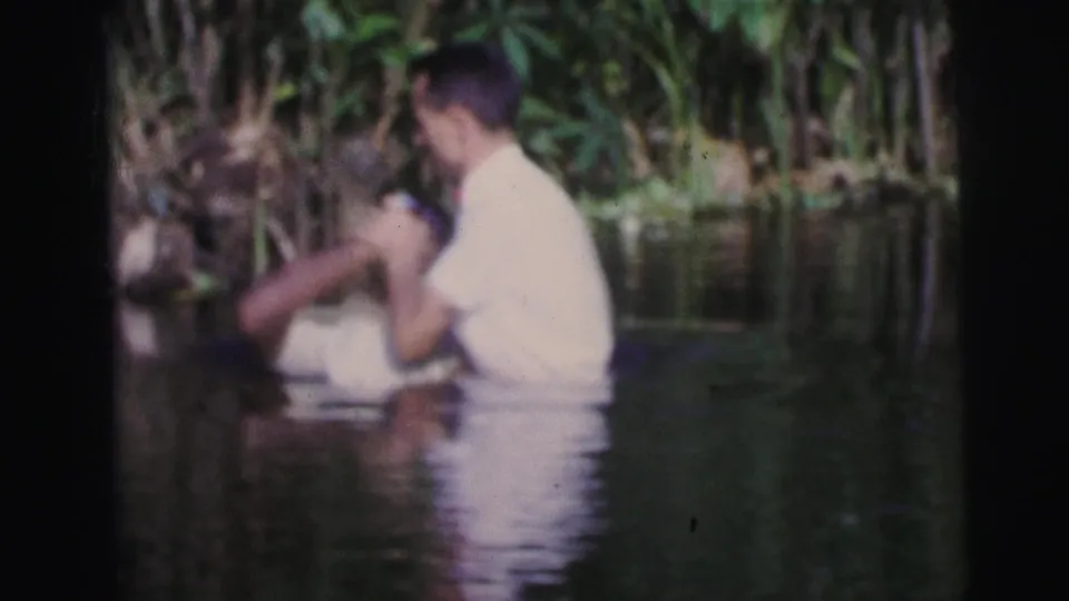 1967 Brazil 8mm frame showing two men in white shirts in a natural water setting during an apparent baptism ritual.